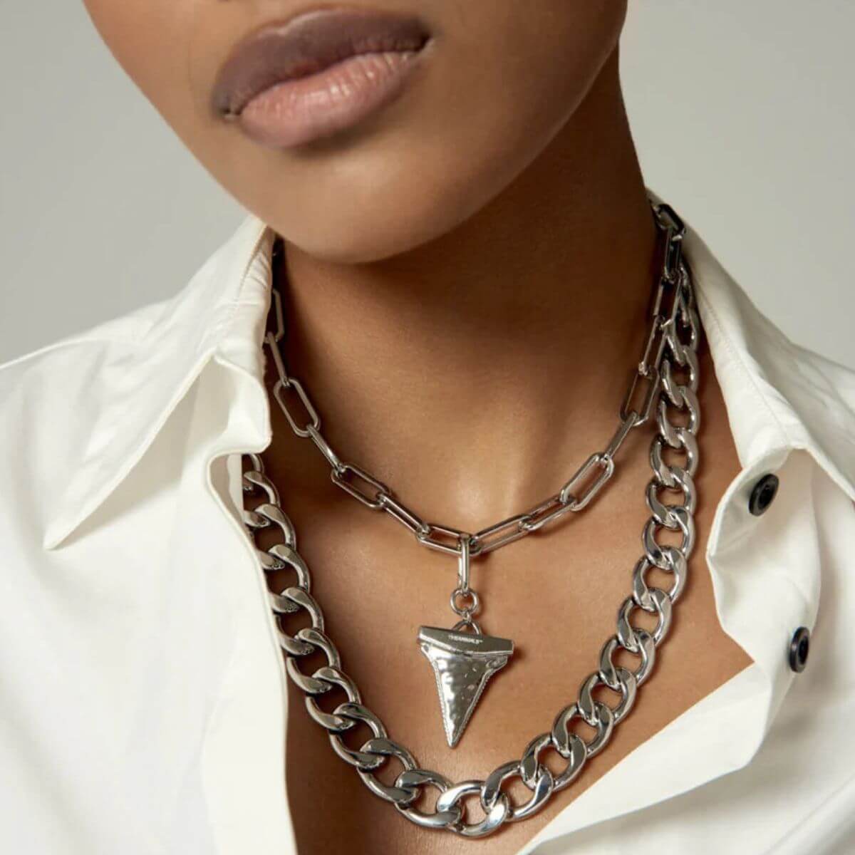 Close-up of woman wearing layered silver necklaces with shark tooth charm by Pendants.
Nahaufnahme einer Frau mit silbernen Ketten im Layering-Look und Haifischzahn-Anhänger von Pendants.
Gros plan d’une femme portant des colliers argentés superposés avec pendentif dent de requin de Pendants.
