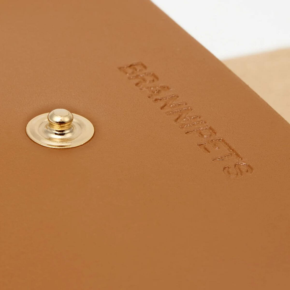 Close-up of the golden button and embossed logo on the tan leather Branni Pets poop bag holder, capturing its elegant craftsmanship. Nahaufnahme des goldenen Druckknopfs und des geprägten Logos auf dem hellbraunen Leder-Kotbeutelhalter von Branni Pets – ein Ausdruck von Eleganz und Qualität. Gros plan sur le bouton doré et le logo embossé du porte-sacs à crottes en cuir camel Branni Pets – une finition élégante et soignée.