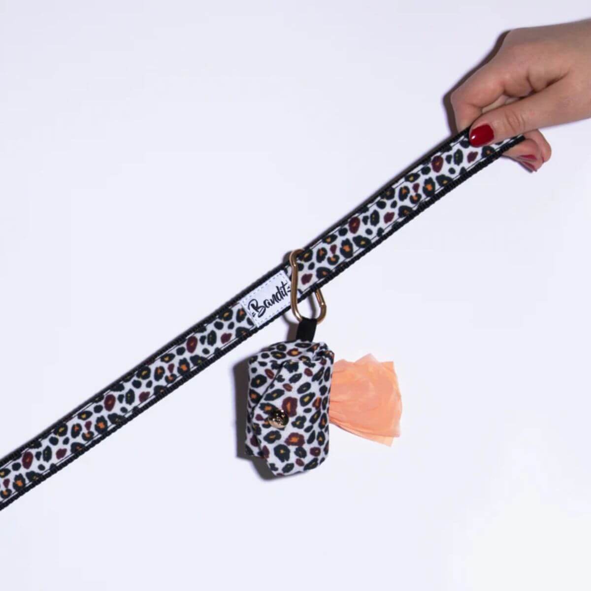 Hand holding a French Bandit leash with matching leopard print poop bag holder and gold clip – bold and coordinated accessory. Hand hält French Bandit Hundeleine mit passendem Kotbeutelhalter im Leopardenmuster und goldenem Clip – auffälliges, abgestimmtes Accessoire. Main tenant une laisse French Bandit avec distributeur de sacs assorti à imprimé léopard et mousqueton doré – accessoire audacieux et assorti.