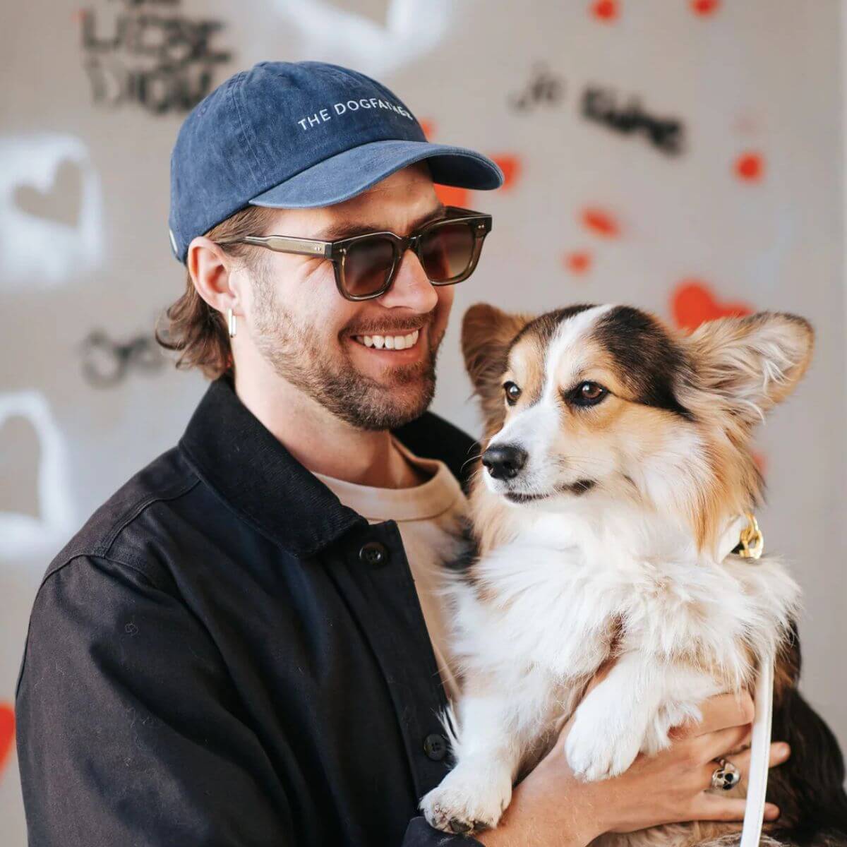 Smiling man wearing a blue washed denim "THE DOGFATHER" cap by Collar of Sweden, holding a fluffy Corgi dog – Homme souriant portant une casquette bleue en denim délavé "THE DOGFATHER" de Collar of Sweden, tenant un chien Corgi à poil long – Lachender Mann mit blauer „THE DOGFATHER“-Kappe aus gewaschenem Denim von Collar of Sweden, hält einen flauschigen Corgi-Hund im Arm.