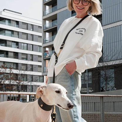 Woman walking a light-colored Greyhound wearing a wide black leather dog collar from Branni Pets, designed for Greyhounds or dogs with sensitive necks. Spaziergang mit einem hellen Windhund, der ein breites schwarzes Lederhalsband von Branni Pets trägt – ideal für Windhunde oder Hunde mit empfindlichem Hals. Femme promenant un lévrier clair avec un collier large en cuir noir signé Branni Pets, conçu pour les lévriers ou les chiens au cou sensible.
