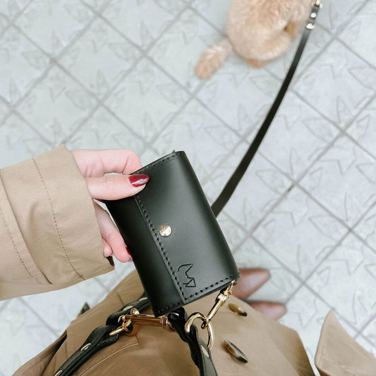Black Branni Pets poop bag holder clipped onto a beige trench coat with matching leash and gold hardware, ready for city walks. Schwarzer Branni Pets Kotbeutelhalter an einem beigen Trenchcoat mit passender Leine und goldenen Details – perfekt für Spaziergänge in der Stadt. Porte-sacs à crottes noir Branni Pets accroché à un trench beige avec une laisse assortie et des finitions dorées – idéal pour les promenades urbaines.