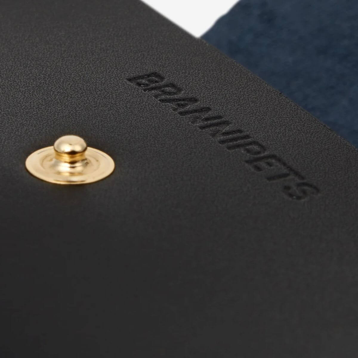 Close-up of the embossed Branni Pets logo on the black vegan leather dog poop bag holder with gold snap button. Nahaufnahme des geprägten Branni Pets Logos auf dem schwarzen Kotbeutelspender aus veganem Leder mit goldfarbenem Druckknopf. Gros plan sur le logo Branni Pets embossé sur le porte-sacs à crottes en cuir vegan noir avec bouton pression doré.