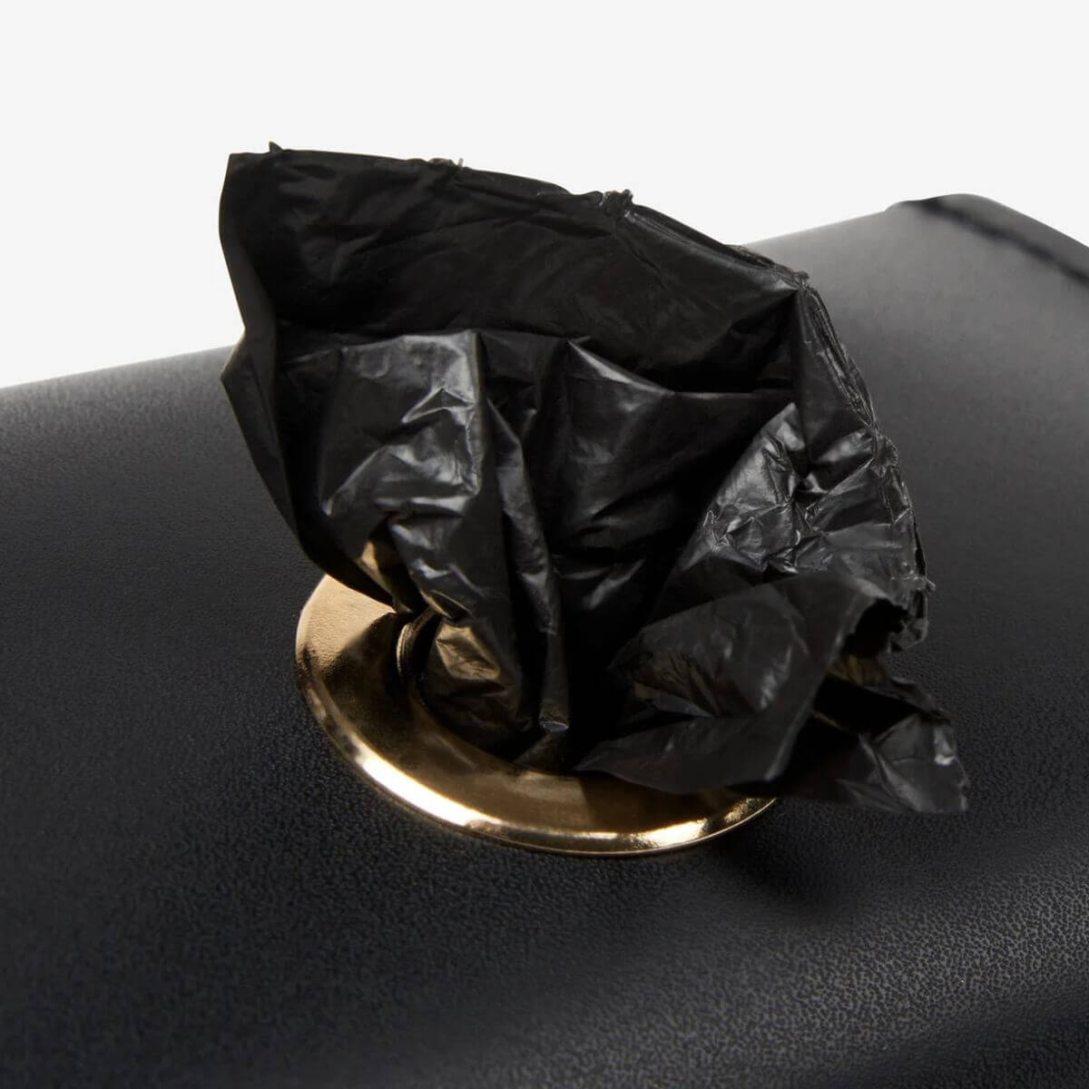 Detail shot of a black waste bag being pulled through the gold-rimmed dispenser opening of the Branni Pets black poop bag holder. Detailaufnahme eines schwarzen Kotbeutels, der durch die goldene Spenderöffnung des schwarzen Branni Pets Kotbeutelhalters gezogen wird. Gros plan d’un sac noir sortant par l’ouverture dorée du distributeur du porte-sacs à crottes noir Branni Pets.