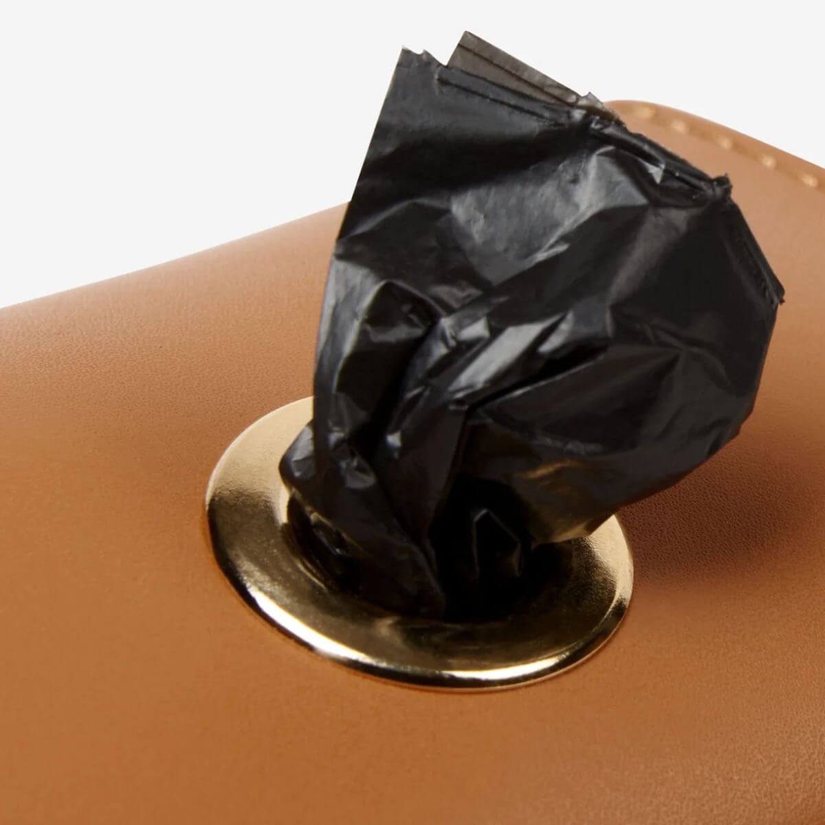 This close-up captures a tan Branni Pets poop bag holder with a black bag peeking through the gold-rimmed opening, ideal for chic dog walks. Nahaufnahme des hellbraunen Branni Pets Kotbeutelhalters mit einem schwarzen Beutel, der durch die goldgerahmte Öffnung ragt – perfekt für stilvolle Hundespaziergänge. Ce gros plan montre le porte-sacs camel Branni Pets avec un sac noir sortant de l’ouverture dorée – idéal pour des promenades chics avec votre chien.