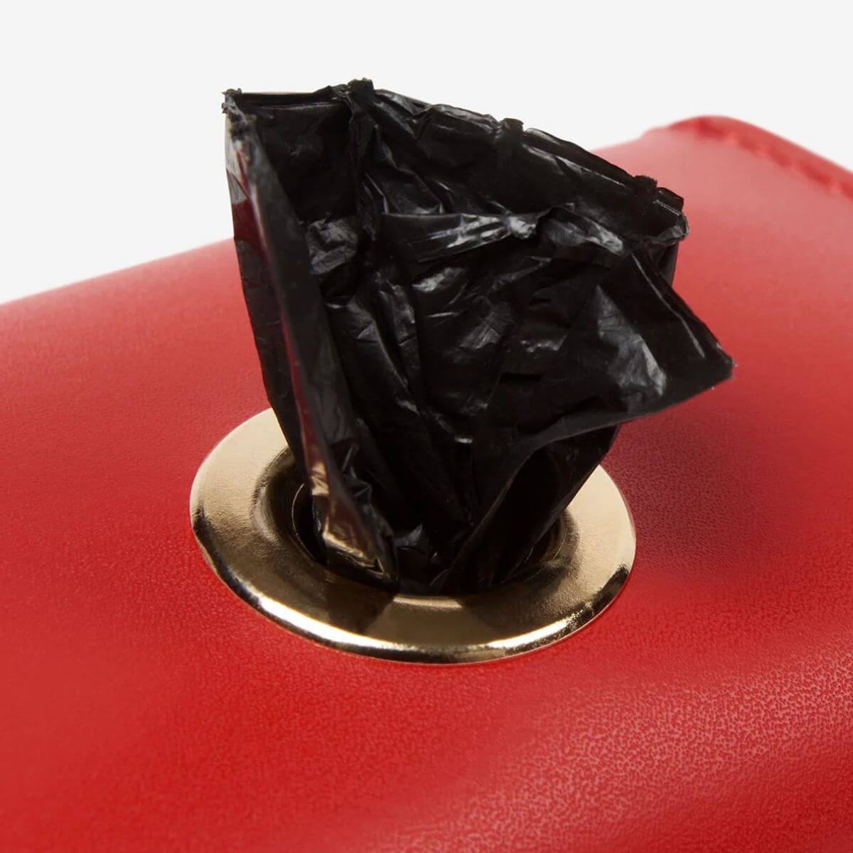 Detail view of Branni Pets red poop bag holder with black bag peeking out through gold-ring opening. Detailansicht des roten Branni Pets Kotbeutelhalters mit herausstehendem schwarzen Beutel durch die goldene Öffnung. Vue de détail du distributeur de sacs rouges Branni Pets avec sac noir sortant par l’anneau doré.