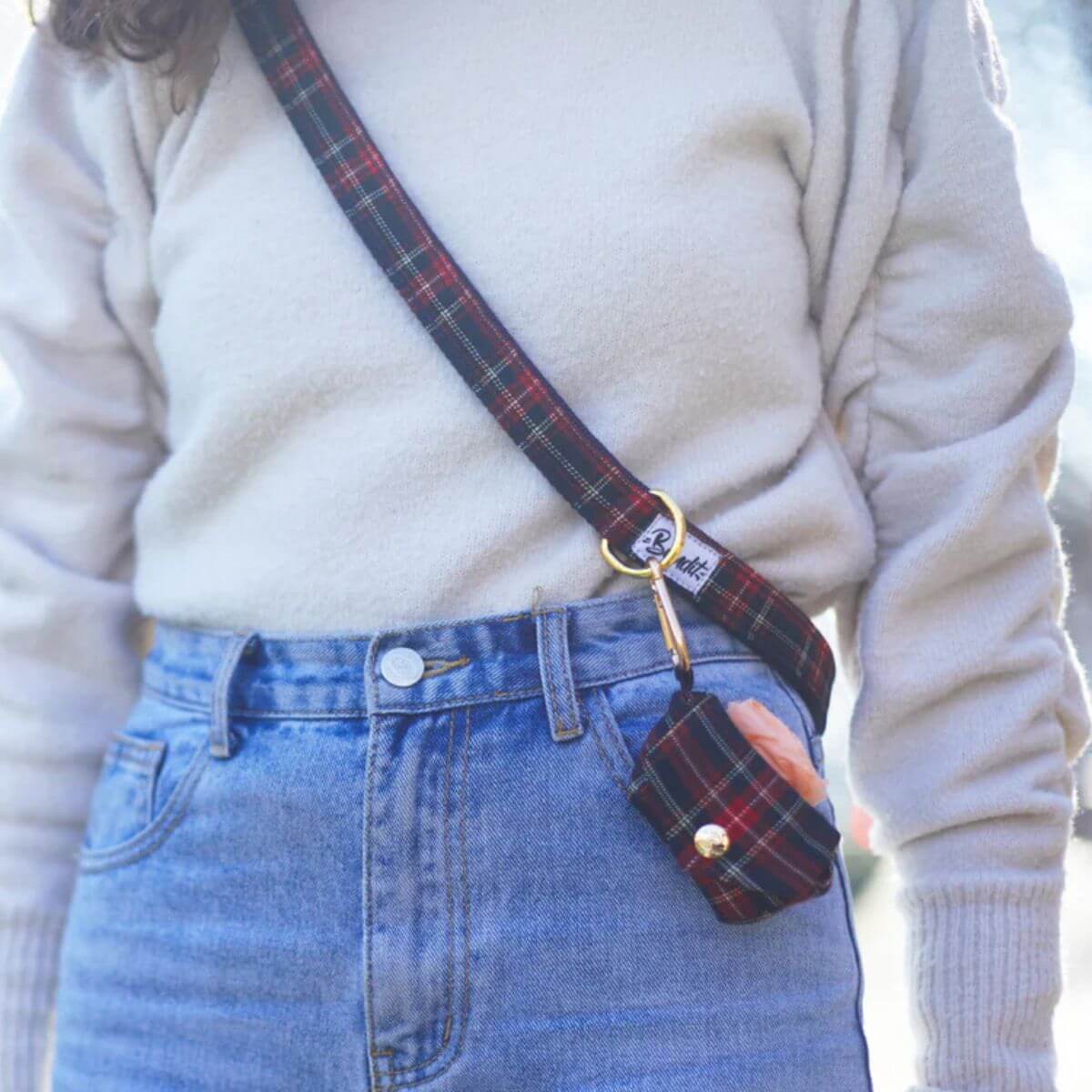 Woman wearing a red tartan French Bandit leash crossbody with attached poop bag holder – functional and fashionable hands-free solution. Frau trägt French Bandit Hundeleine im roten Tartan als Crossbody mit angehängtem Kotbeutelhalter – modisch und freihändig unterwegs. Femme portant la laisse tartan rouge de French Bandit en bandoulière avec porte-sac attaché – pratique et tendance pour les balades mains libres.