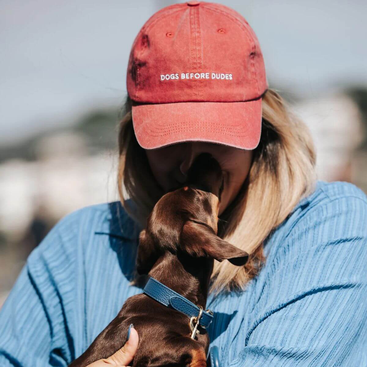 Woman in blue shirt wearing "DOGS BEFORE DUDES" cap from Collar of Sweden, cuddling dachshund puppy. Frau im blauen Hemd mit "DOGS BEFORE DUDES"-Cap von Collar of Sweden, kuschelt mit Dackelwelpen. Femme en chemise bleue portant une casquette "DOGS BEFORE DUDES" de Collar of Sweden, câlinant un chiot teckel.