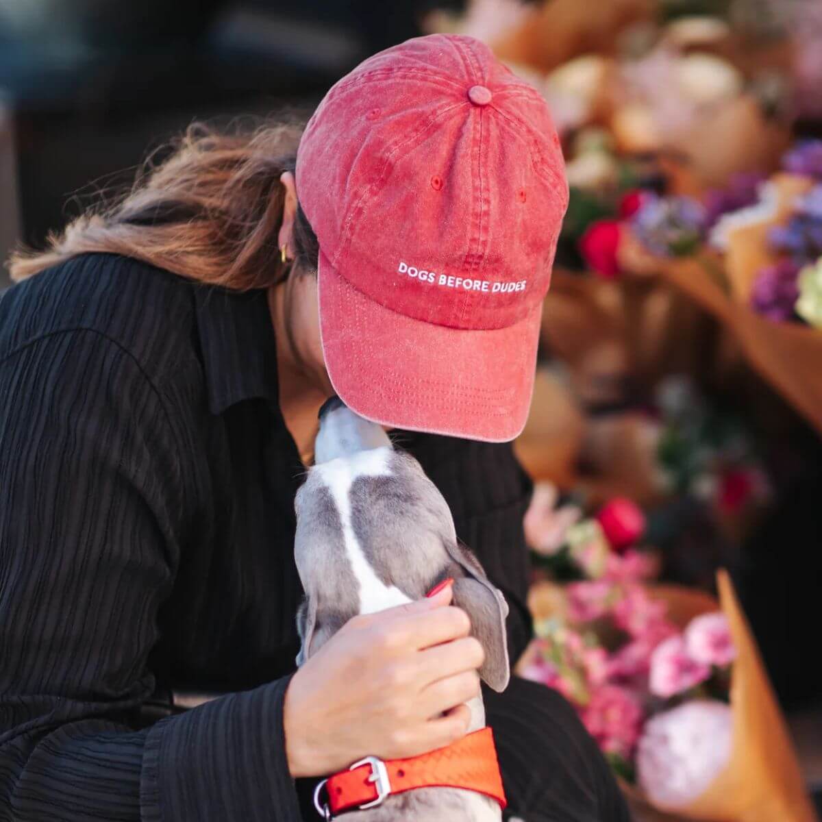 Woman wearing "DOGS BEFORE DUDES" red cap from Collar of Sweden, embracing her whippet among flowers. Frau mit roter "DOGS BEFORE DUDES"-Cap von Collar of Sweden, umarmt ihren Whippet zwischen Blumen. Femme avec casquette rouge "DOGS BEFORE DUDES" de Collar of Sweden, enlaçant son whippet parmi les fleurs.