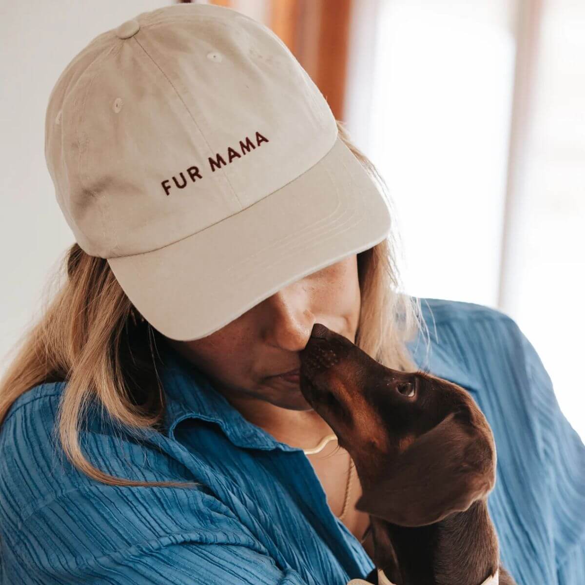 Woman wearing a washed beige denim cap from Collar of Sweden with "FUR MAMA" embroidered on the front, kissing her dachshund – Femme portant une casquette de Collar of Sweden en jean beige délavé avec l'inscription "FUR MAMA" brodée à l'avant, embrassant son teckel – Frau mit beigefarbener, gewaschener Denim-Kappe von Collar of Sweden und „FUR MAMA“-Stickerei auf der Vorderseite, küsst ihren Dackel.