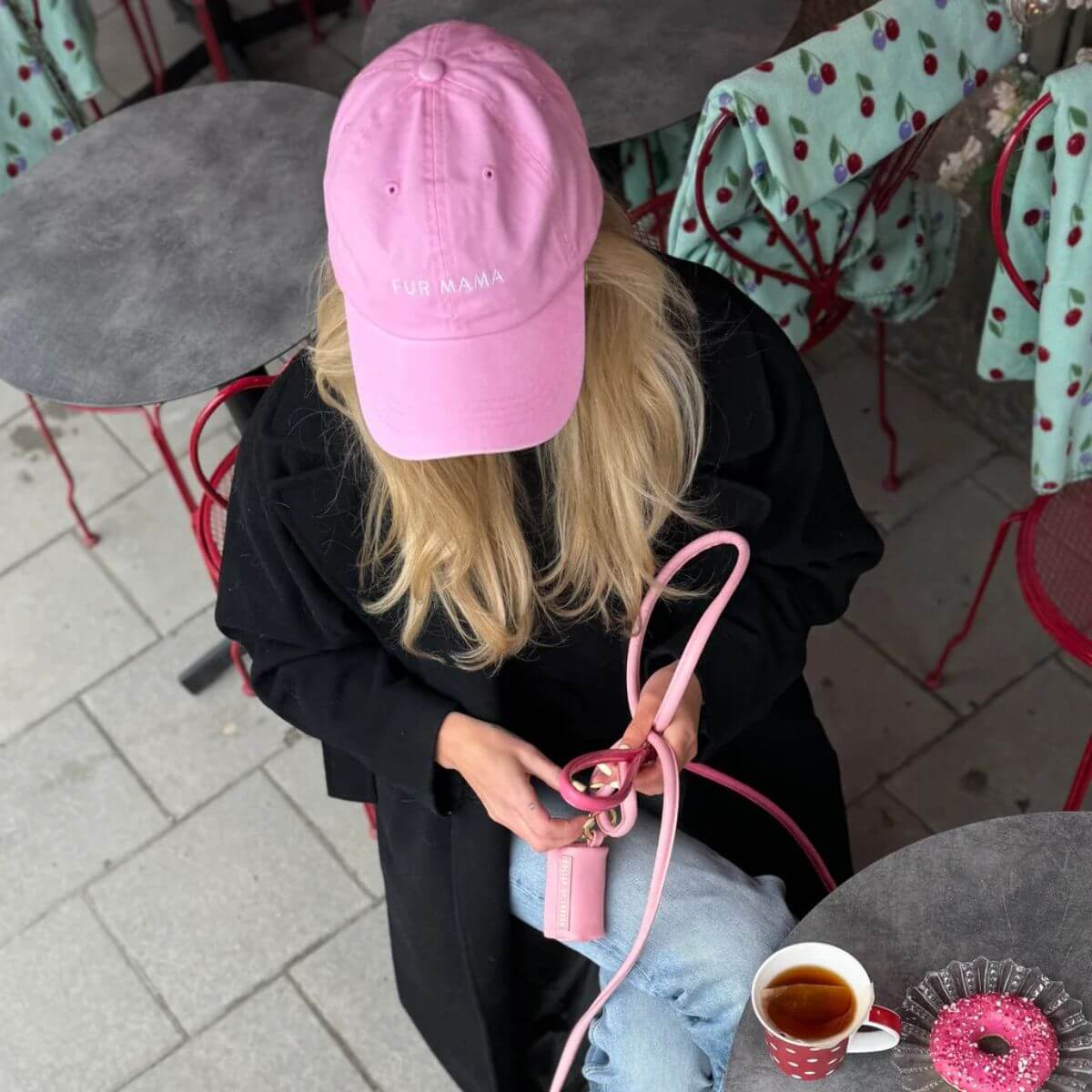Woman with a pink "FUR MAMA" baseball cap from Collar of Sweden drinking a coffee at a café – Femme avec une casquette rose "FUR MAMA" de Collar of Sweden boit un café assise à une terrasse – Frau mit rosa „FUR MAMA“-Kappe von Collar of Sweden trinkt einen Kaffee im Strassencafé.