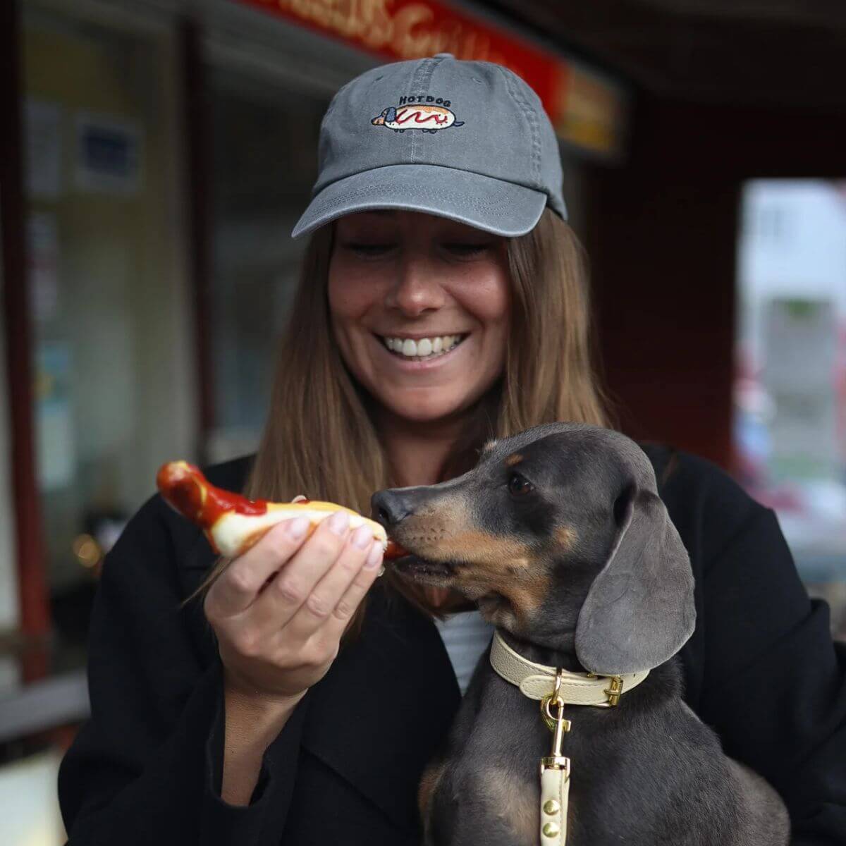 Smiling woman wearing a washed grey “Hot Dog” cap by Collar of Sweden, holding her Dachshund and feeding it a hot dog – designed for dachshund owners and lovers – Femme souriante portant une casquette grise délavée “Hot Dog” de Collar of Sweden, donnant un hot-dog à son teckel – conçue pour les propriétaires et amoureux des teckels – Lachende Frau mit grauer “Hot Dog”-Kappe aus gewaschenem Denim von Collar of Sweden, füttert ihren Dackel mit einem Hotdog – für Dackelbesitzer und -liebhaber.