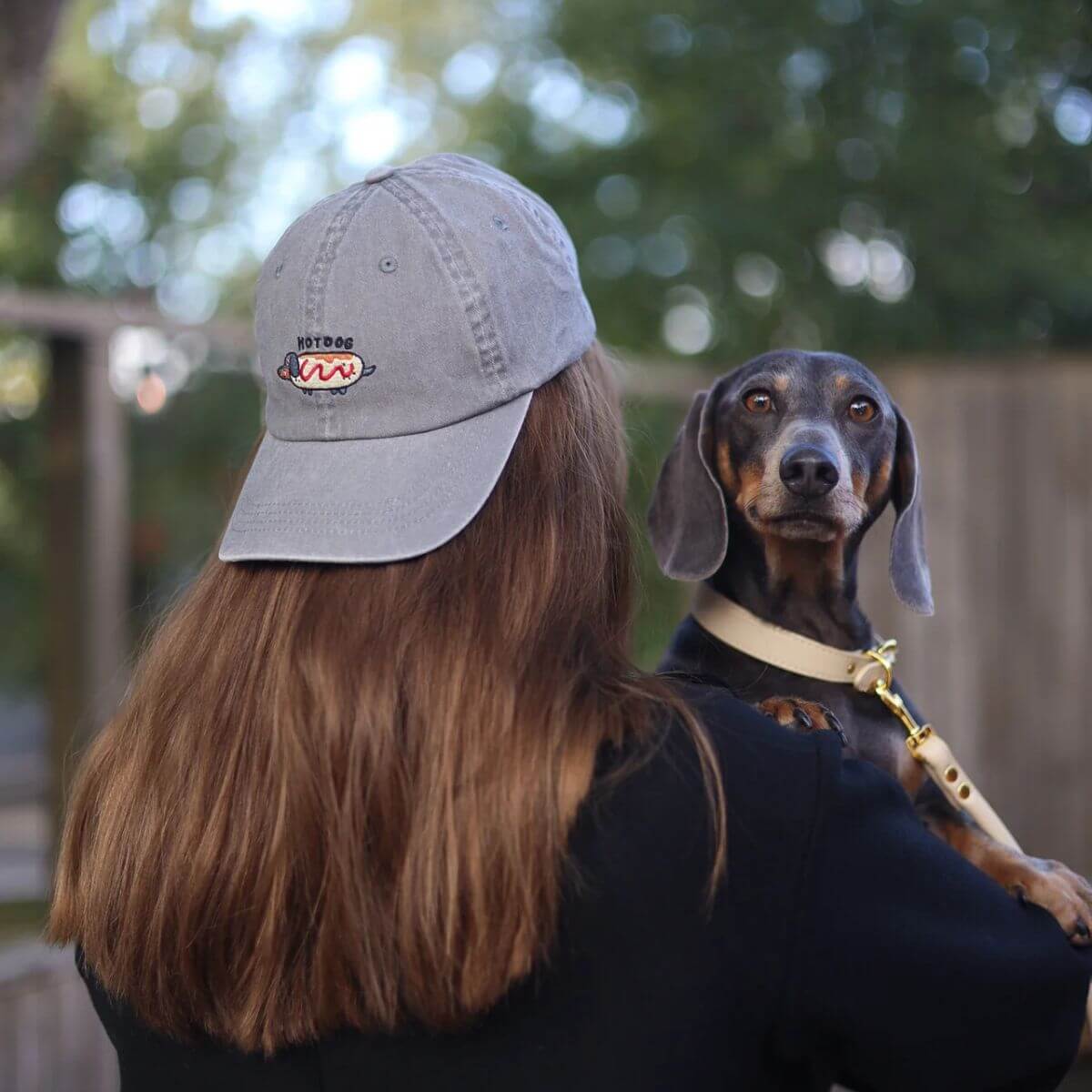 Woman wearing a washed grey denim “Hot Dog” cap by Collar of Sweden, holding a Dachshund – designed for dachshund owners and lovers – Femme portant une casquette en denim gris délavé “Hot Dog” de Collar of Sweden, tenant un teckel – conçue pour les propriétaires et amoureux des teckels – Frau mit grauer, gewaschener Denim-Kappe “Hot Dog” von Collar of Sweden, hält einen Dackel im Arm – für Dackelbesitzer und -liebhaber.